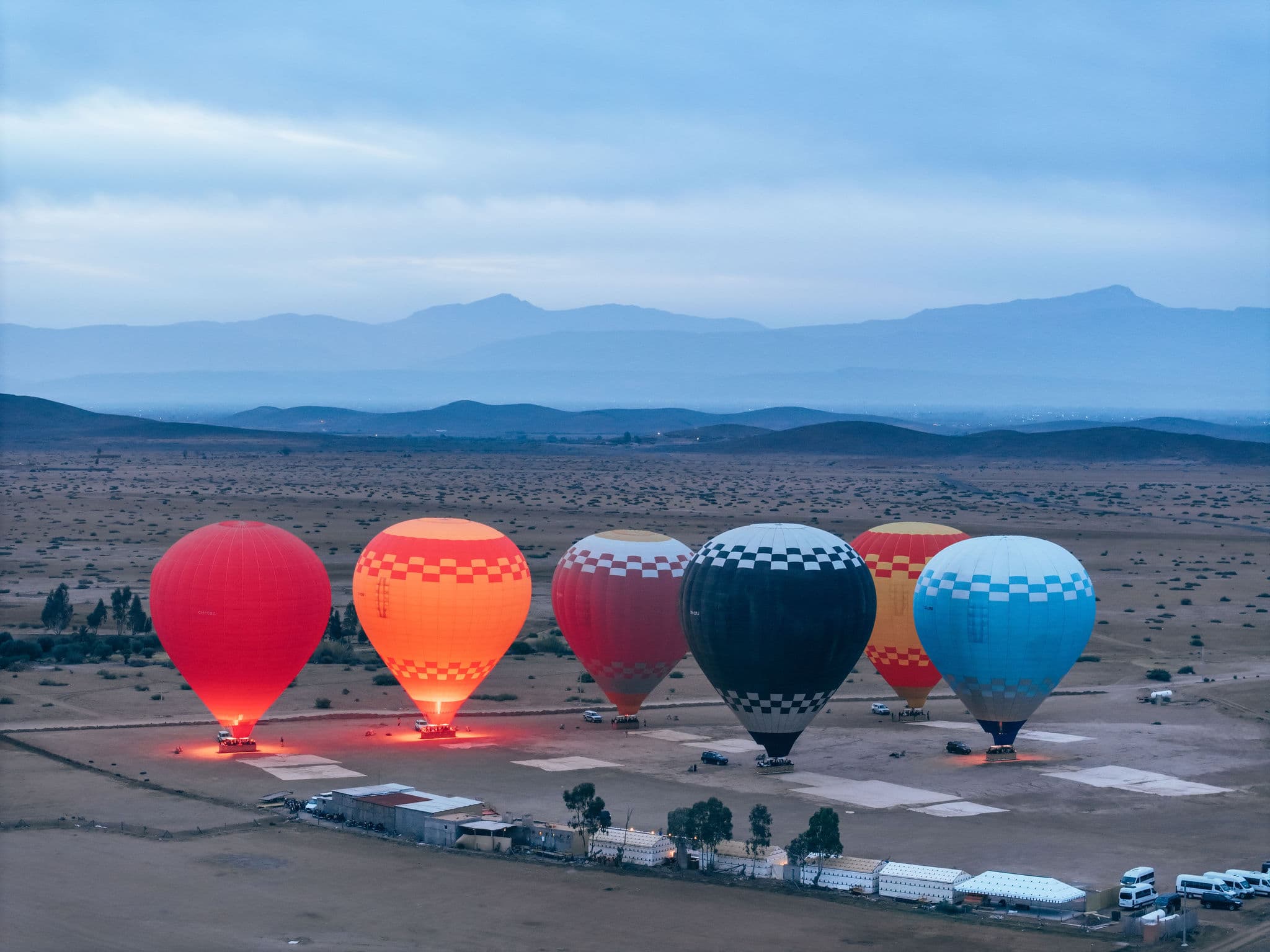 Line of glowing hot air balloons tethered on ground against dusky desert and mountains.